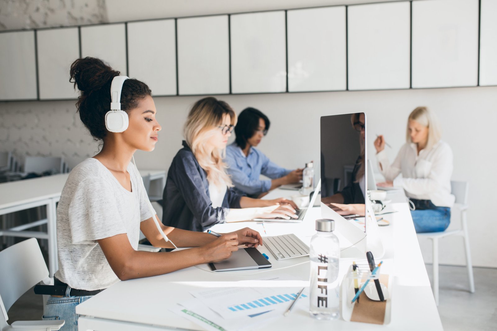 Concentrated african female web-designer using graphics tablet while her colleagues writing reports. Indoor portrait of programmers of international company spending time together at workplace.